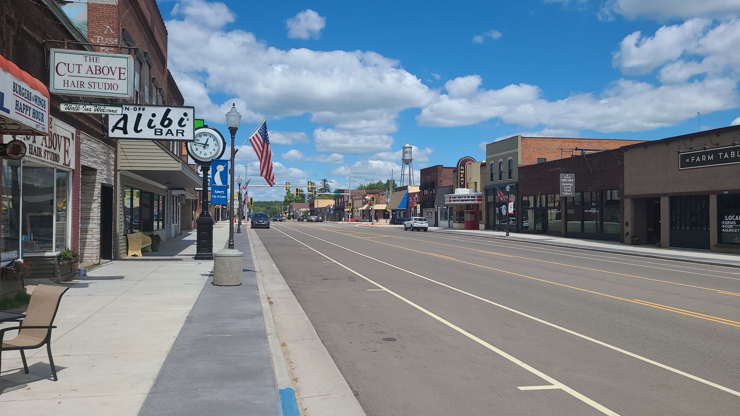 Downtown Amery in summer, a row of brick storefronts along a wide street under a blue sky with scattered clouds.