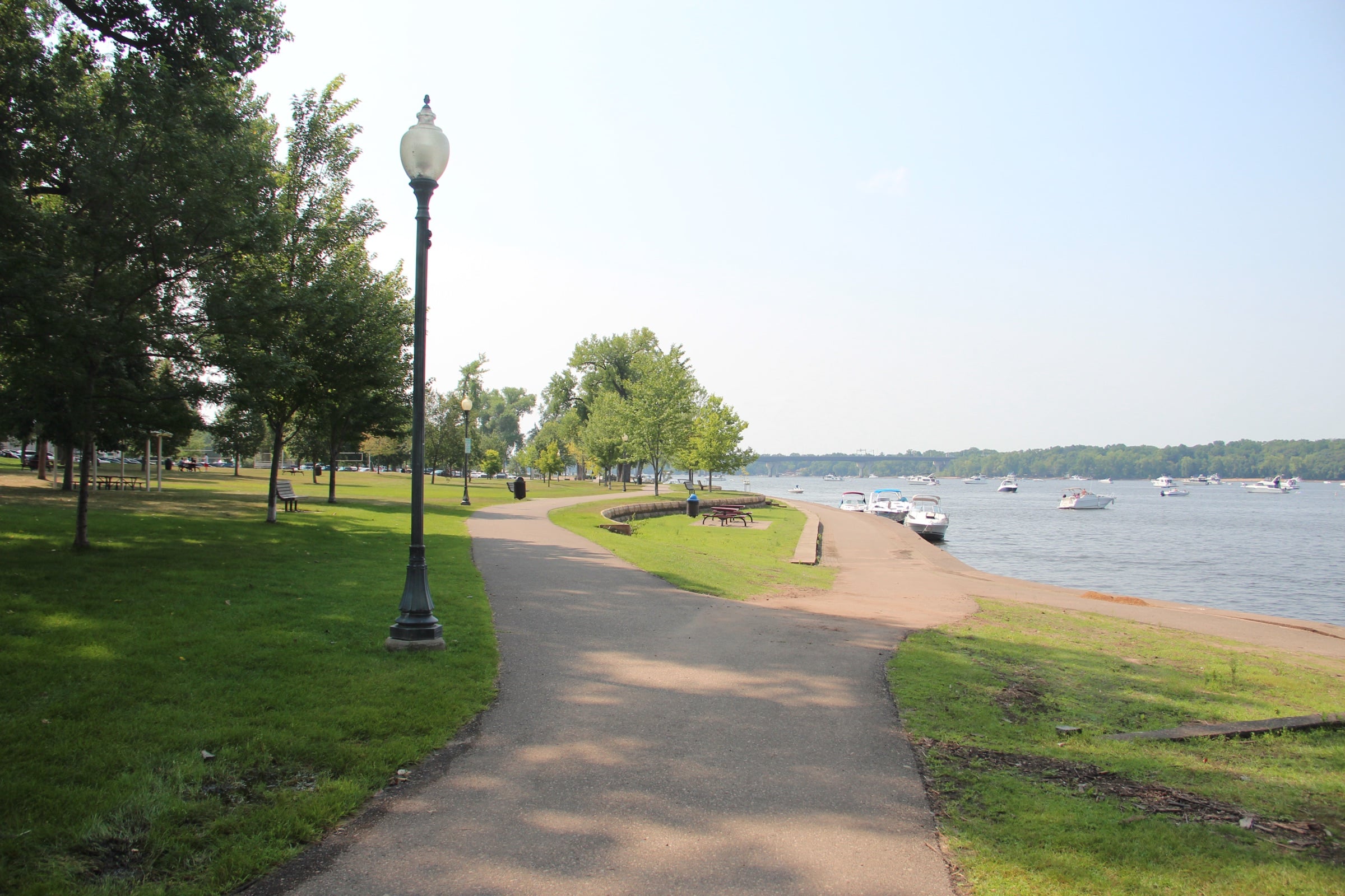 A paved path through Lakefront Park in Hudson beside the St. Croix River, with shade trees and boats moored offshore.