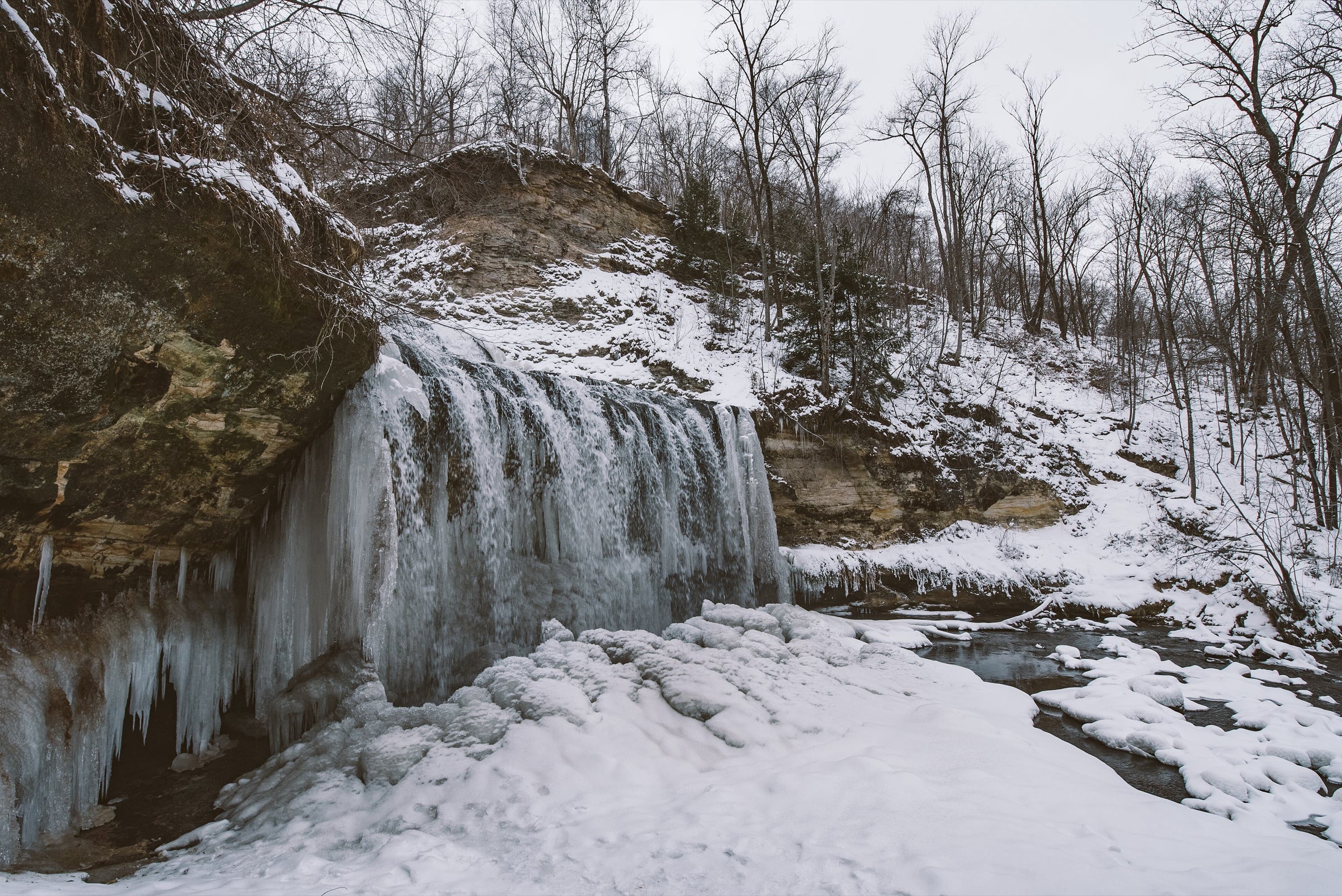 Cascade Falls in Osceola in winter, partly frozen over a rock ledge, with snow in the glen and bare trees on the bluff behind.